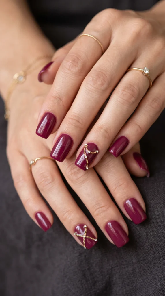Square deep berry nails with draped gold chain accent nail art and rhinestone details on ring finger